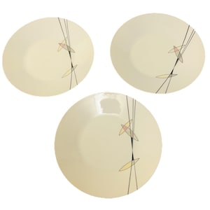 Op de afbeelding: Drie crèmekleurige keramische borden met een modern design uit het midden van de eeuw. Elk bord heeft een zwart lijnenontwerp met gele en roze geometrische vormen. De borden zijn rond en hebben een eenvoudige, elegante uitstraling.