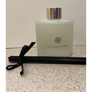 May include: A frosted glass diffuser bottle with a silver cap, labeled "SUPPER CLUB" with a tree logo. Black reeds are tied with a black ribbon. The bottle is square and contains a light-colored liquid.