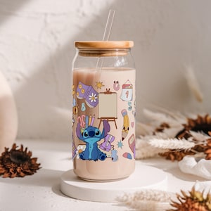 May include: A clear glass tumbler with a wooden lid and a straw, filled with a light-colored beverage. The tumbler is decorated with a colorful cartoon design featuring Stitch, art supplies, and a calendar. The tumbler sits on a white pedestal.