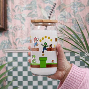 May include: A clear glass drinking cup with a wooden lid and a metal straw. The cup is decorated with a colorful Super Mario Bros. design featuring Mario, Luigi, Yoshi, and other characters. The cup is filled with a white liquid.