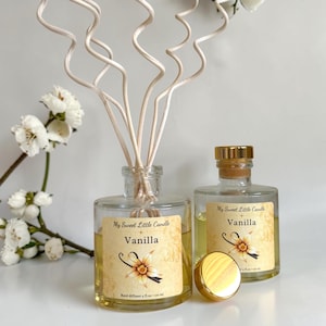 May include: Two clear glass vanilla reed diffusers with gold lids and labels. The labels read "My Sweet Little Candle" and "Vanilla" with a vanilla flower graphic. The bottles are filled with a yellow liquid. Reed sticks are inserted into one bottle.
