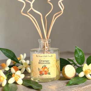 May include: A clear glass reed diffuser bottle with a label that reads "Orange Blossom" and "My Sweet Little Candle." The bottle contains a yellow liquid and is surrounded by flowers and leaves. The diffuser is 4 fl oz / 120 ml.