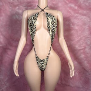 Op de afbeelding: Een badpak met slangenprint op een mannequin. De haltertop en de string-stijl broek zijn verbonden door dunne zwarte bandjes. De print heeft tinten beige, bruin en zwart, waardoor een realistisch slangenpatroon ontstaat. De mannequin staat tegen een roze achtergrond.