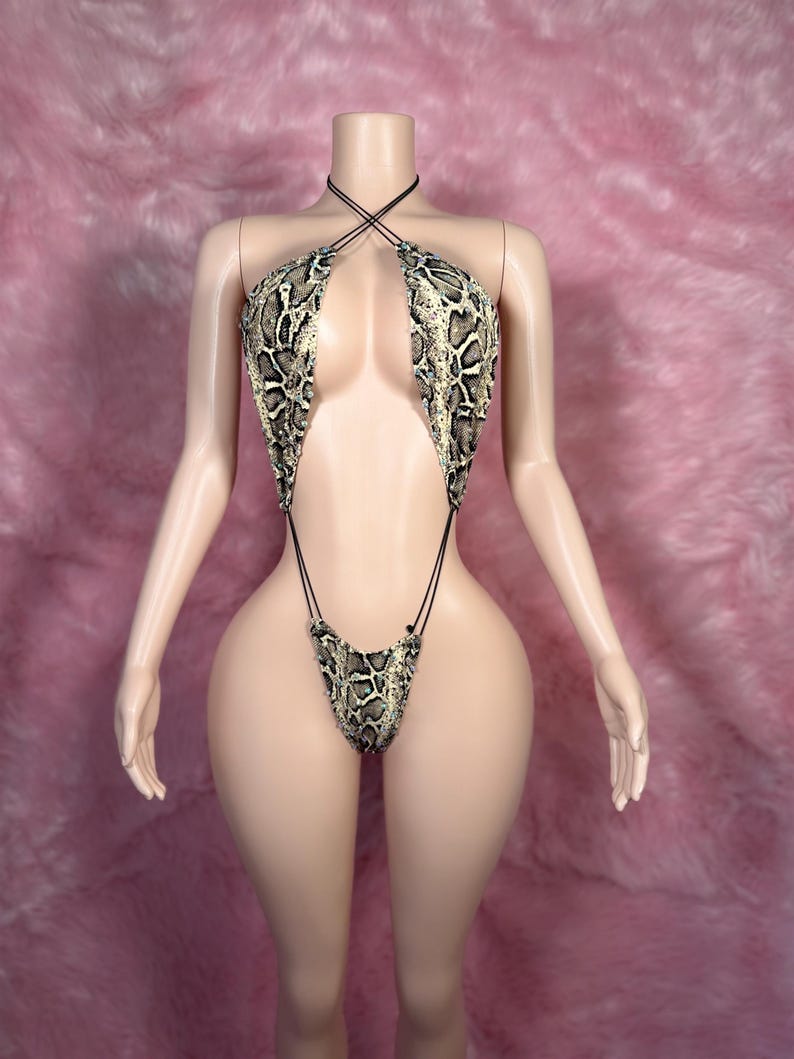 Op de afbeelding: Een badpak met slangenprint op een mannequin. De haltertop en de string-stijl broek zijn verbonden door dunne zwarte bandjes. De print heeft tinten beige, bruin en zwart, waardoor een realistisch slangenpatroon ontstaat. De mannequin staat tegen een roze achtergrond.