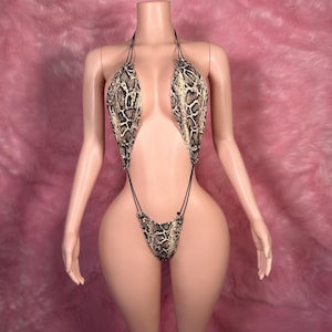 Peut inclure: Un maillot de bain à imprimé peau de serpent avec un haut dos nu et un bas en string. Le maillot de bain présente un motif marron, beige et noir. Le vêtement est présenté sur un mannequin avec un fond rose.