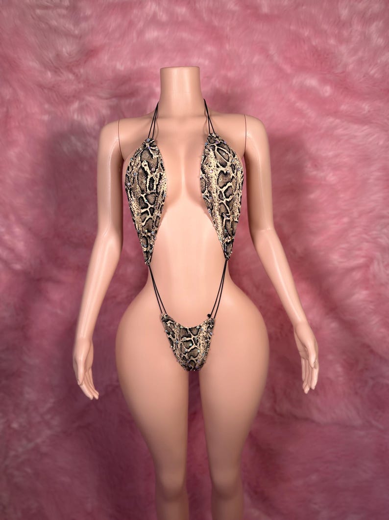 Op de afbeelding: Een badpak met slangenprint met een haltertop en een stringbroekje. Het badpak heeft een bruin, beige en zwart patroon. Het kledingstuk wordt gepresenteerd op een mannequin met een roze achtergrond.
