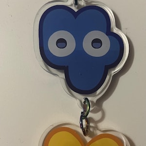 May include: A blue and yellow cartoon character keychain with large eyes. The keychain is made of clear acrylic and has a metal ring for attaching to a bag or keys.