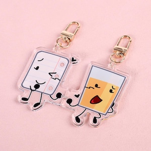 May include: Two acrylic keychains featuring cartoon characters. The first keychain is a white notepad with a red heart and a black face. The second keychain is a yellow glass with a black face and a red mouth. Both keychains have gold key rings.