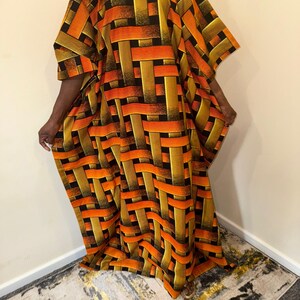 Könnte beinhalten: Ein bodenlanges Kaftan-Kleid mit einem lebendigen Webmuster in Orange-, Gelb- und Schwarztönen. Das Kleidungsstück hat einen Rundhalsausschnitt, weite kurze Ärmel und eine lockere, fließende Silhouette mit einem sich wiederholenden geometrischen Flechtmuster.