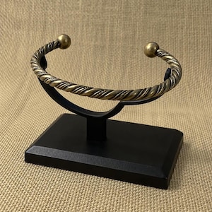 Puede incluir: Un brazalete de metal en tono latón con un diseño de cuerda trenzada y una cuenta esférica lisa en cada extremo abierto. El brazalete se exhibe sobre un soporte negro que tiene una base rectangular y un brazo de soporte curvo para la joya.