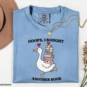 Può includere: T-shirt in denim blu chiaro lavato con una grafica di un'anatra bianca che trasporta una pila di libri. Il testo recita "OOOPS, I BOUGHT ANOTHER BOOK". Una collana a stella dorata è drappeggiata sulla maglietta.