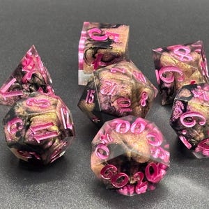 May include: A set of seven polyhedral dice made of clear resin with swirling black, gold, and purple metallic inclusions. Each die features recessed numbers painted in a vibrant, glossy hot pink, providing high contrast against the dark, swirling interior.