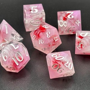 May include: A set of seven polyhedral dice for tabletop games. The dice are a translucent pink with red and white floral inclusions. Each die has silver numbering. The set includes a D4, D6, D8, D10, D12, D20, and a percentile die.