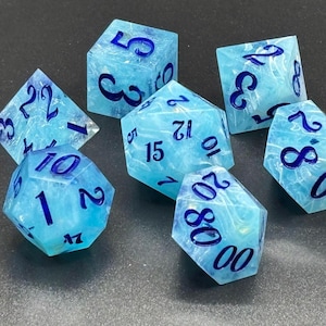 May include: A set of seven polyhedral dice featuring a translucent light blue and white marbled pattern with dark blue ink numbering. The collection includes a d4, d6, d8, d10, d12, d20, and a percentile die, all displaying clear, legible numerals.