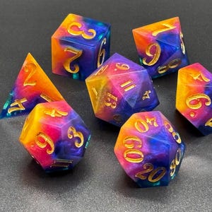 Puede incluir: Un juego de siete dados poliédricos con una mezcla de resina en tonos naranja vibrante, rosa y azul profundo. Cada dado tiene números grabados con un acabado en oro metálico, incluyendo un d20, d12, d10, d8, d6, d4 y un dado de porcentaje.