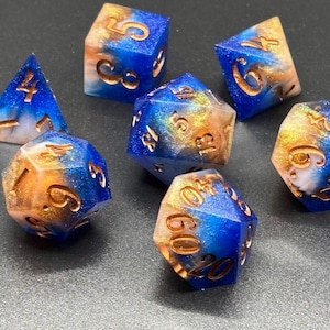 May include: A set of seven polyhedral dice featuring a swirling blend of deep blue, bright orange, and white resin with a glittery finish. Each die has engraved numbers filled with a metallic copper color, including a d4, d6, d8, two d10s, a d12, and a d20.