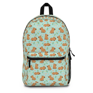 May include: A light blue backpack featuring a repeating pattern of brown capybaras, small yellow flowers, and green leaves. The backpack has a black top handle, a front zippered pocket, and a black zipper closure.