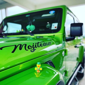 May include: Bright green Jeep with the word "Mojiteaux" in black script on the hood. A small rubber ducky wearing a green hat sits on the hood. The vehicle has black side mirrors and a black front grill.