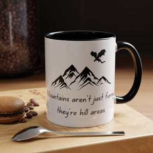 Puede incluir: Taza de cerámica blanca con borde y asa negros. La taza presenta una silueta negra de montañas y un dragón volando sobre ellas. El texto "Mountains aren't just funny they're hill areas." está impreso en la taza.