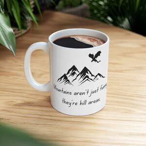 Puede incluir: Taza de café de cerámica blanca con una cordillera de montañas negra y una silueta de dragón. La taza dice "Mountains aren't just funny, they're hill areas."