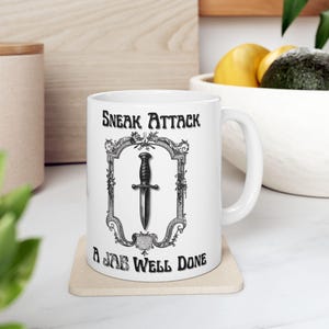 May include: White ceramic mug with a black and white illustration of a dagger in a decorative frame. The text "Sneak Attack" and "A Jab Well Done" are printed on the mug.