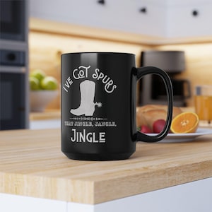May include: Black coffee mug with white text that reads "I've Got Spurs That Jingle, Jangle, Jingle". The text is surrounded by a decorative border. The mug has a white image of a cowboy boot with a spur.