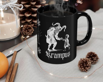 Puede incluir: Taza de café de cerámica negra con una ilustración blanca de Krampus, una criatura mítica, sosteniendo la mano de un niño. El texto "Krampus" está escrito debajo de la ilustración.