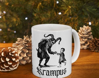 Puede incluir: Taza de cerámica blanca con una ilustración en blanco y negro de Krampus y un niño. La palabra "Krampus" está escrita en una fuente gótica. La taza tiene un asa curva. Decoración con piñas.