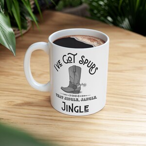May include: White ceramic coffee mug with a black handle. The mug has a black and white illustration of a cowboy boot with spurs and the text "I've Got Spurs That Jingle, Jangle, Jingle".