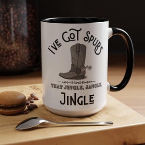 Könnte beinhalten: Weiße Keramik-Kaffeetasse mit schwarzem Rand und Henkel. Die Tasse zeigt einen braunen Cowboy-Stiefel mit Sporen und dem Text "I've Got Spurs That Jingle, Jangle, Jingle".
