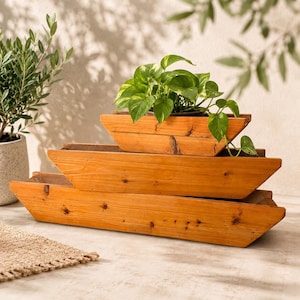 May include: Three stacked, boat-shaped wooden planters with a natural light brown finish and visible wood grain. The top planter contains a green pothos plant with variegated leaves. The planters have angled sides and a long, rectangular base.
