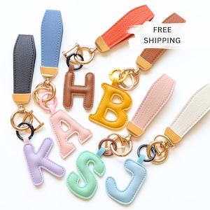 May include: A collection of colorful initial keychains. Each keychain features a letter in various pastel colors, attached to a wrist strap with a gold-tone clasp. The letters are in brown, yellow, pink, green, blue, and purple.