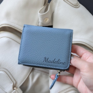 May include: A dusty blue, pebbled leather bifold wallet featuring the name Madeline embossed in a dark script font on the front. The wallet includes a side zipper closure with a matching blue leather pull tab and visible stitching along the edges.