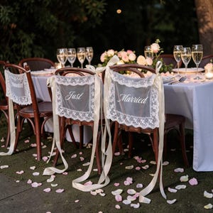 Könnte beinhalten: Hochzeitsempfang mit Holzstühlen, die mit Spitzen-Stuhlhussen geschmückt sind. Auf den Hussen steht "Just Married" und "Bridal". Der Tisch ist mit Sektgläsern, Blumen und einer weißen Tischdecke gedeckt. Rosa Rosenblätter sind auf dem Boden verstreut.
