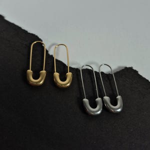 Peut inclure: Quatre boucles d'oreilles en forme d'épingle à nourrice. Deux sont dorées et deux sont argentées. Les boucles d'oreilles sont disposées par paires, chacune ayant un design similaire. Le fond est un mélange de noir et de blanc.