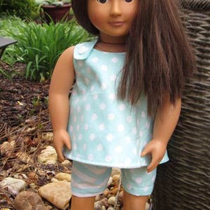 Puede incluir: Una mu&ntilde;eca con el pelo largo y casta&ntilde;o que lleva un vestido sin mangas de lunares blancos sobre fondo azul claro y pantalones cortos de rayas blancas y azules. La mu&ntilde;eca est&aacute; de pie sobre un lecho de peque&ntilde;as piedras.