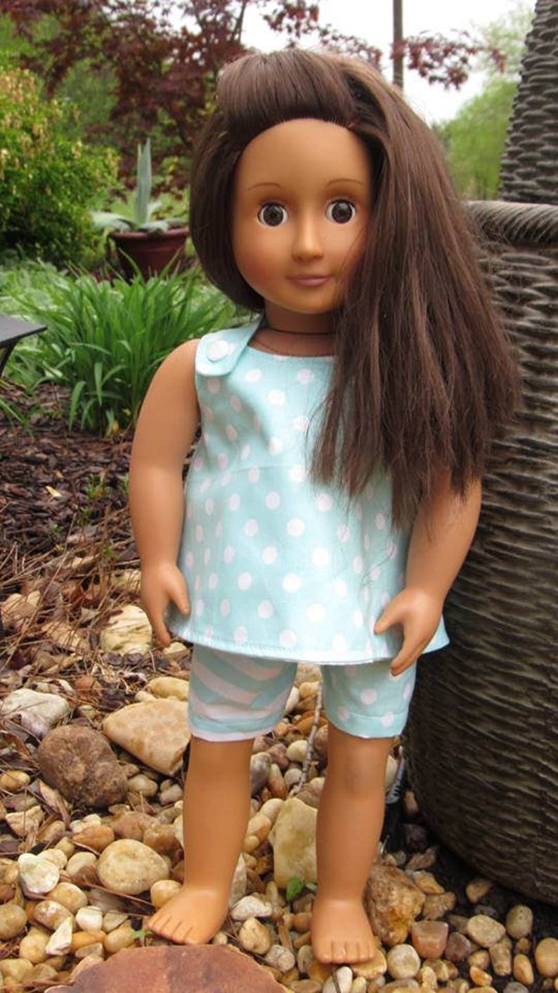 Puede incluir: Una mu&ntilde;eca con el pelo largo y casta&ntilde;o que lleva un vestido sin mangas de lunares blancos sobre fondo azul claro y pantalones cortos de rayas blancas y azules. La mu&ntilde;eca est&aacute; de pie sobre un lecho de peque&ntilde;as piedras.