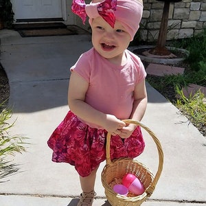 May include: A young child wearing a pink shirt and a floral skirt holds a wicker basket with pink plastic Easter eggs. The child is wearing a pink headband with a bow.
