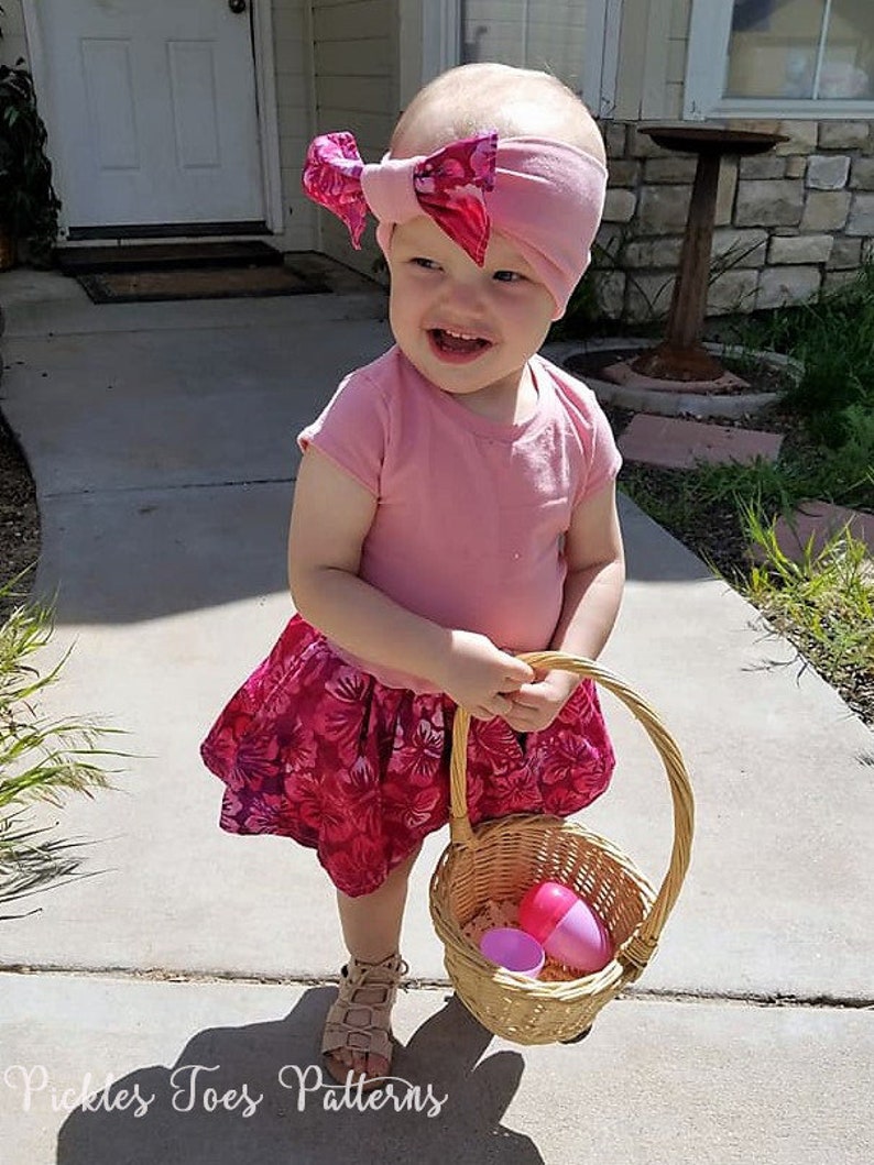 May include: A young child wearing a pink shirt and a floral skirt holds a wicker basket with pink plastic Easter eggs. The child is wearing a pink headband with a bow.