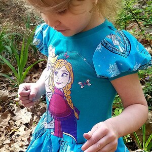 May include: A young girl wearing a blue dress with a printed image of Elsa and Anna from the movie Frozen. The dress has a ruffled skirt and puffed sleeves. The girl is standing in a wooded area with leaves on the ground.