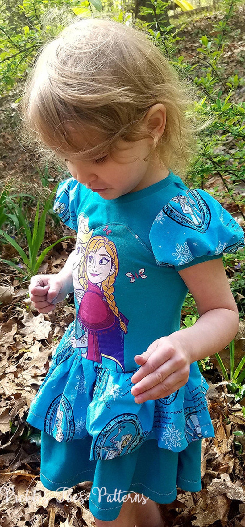 May include: A young girl wearing a blue dress with a printed image of Elsa and Anna from the movie Frozen. The dress has a ruffled skirt and puffed sleeves. The girl is standing in a wooded area with leaves on the ground.