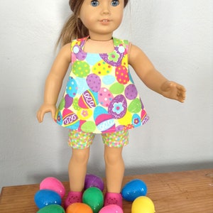 Puede incluir: Una mu&ntilde;eca con un vestido y pantalones cortos con estampado de huevos de Pascua coloridos se encuentra de pie sobre una superficie de madera rodeada de huevos de Pascua de pl&aacute;stico en varios colores.