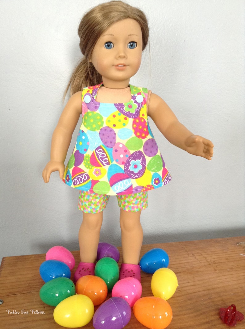 Puede incluir: Una mu&ntilde;eca con un vestido y pantalones cortos con estampado de huevos de Pascua coloridos se encuentra de pie sobre una superficie de madera rodeada de huevos de Pascua de pl&aacute;stico en varios colores.