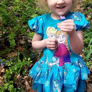 May include: A young girl wearing a blue dress with a print of two girls from the movie Frozen. The dress has a ruffled skirt and a white snowflake design. The girl is holding a purple flower.