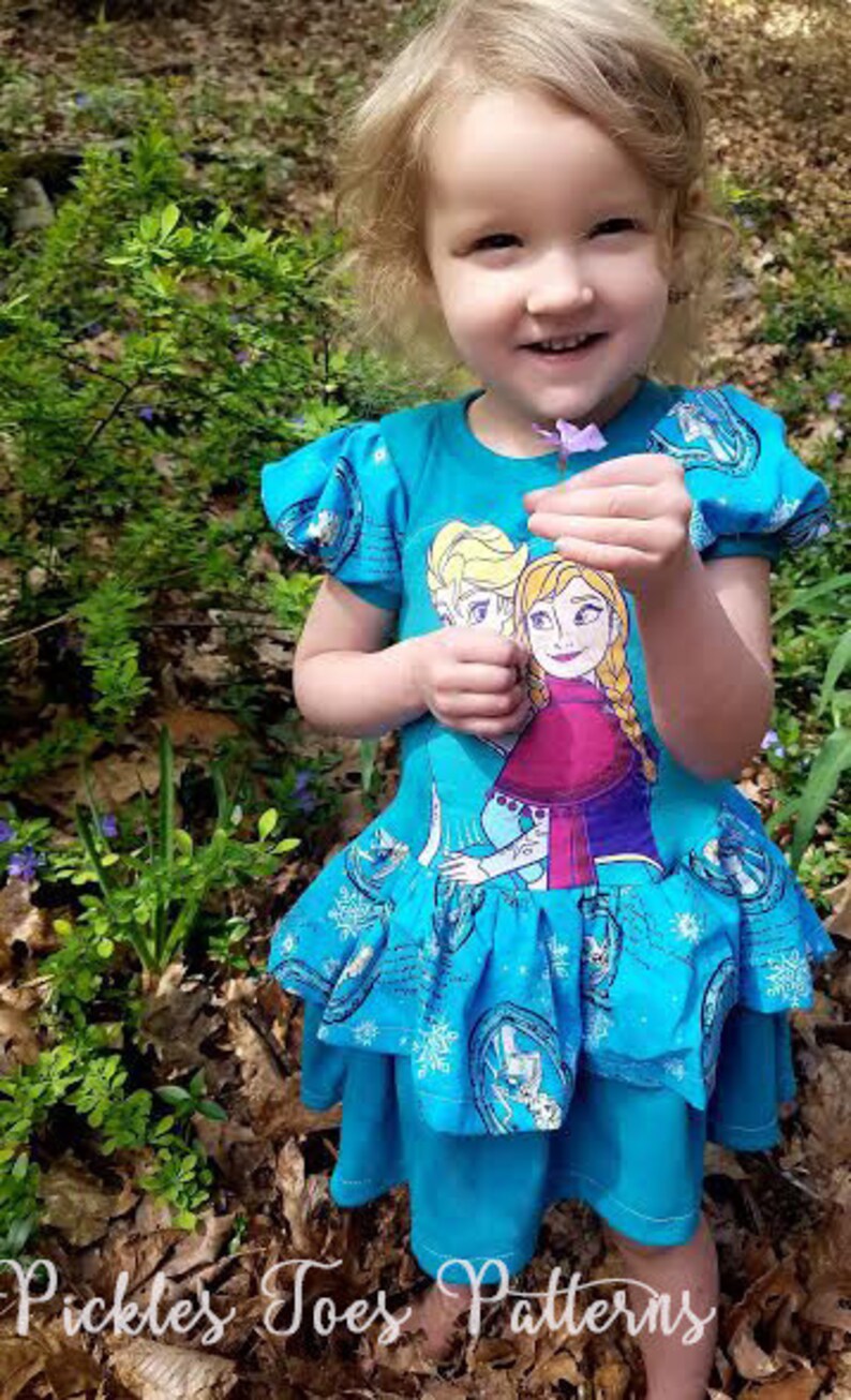 May include: A young girl wearing a blue dress with a print of two girls from the movie Frozen. The dress has a ruffled skirt and a white snowflake design. The girl is holding a purple flower.