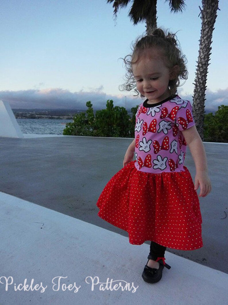 May include: A young girl wearing a pink and red dress with a Minnie Mouse print. The dress has a red polka dot skirt and short sleeves. The girl is standing on a concrete surface with a body of water in the background.