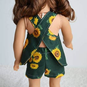 Puede incluir: Una mu&ntilde;eca con un vestido de verano de estampado de girasol verde y amarillo con espalda cruzada. El vestido tiene un dobladillo con volantes y se combina con pantalones cortos a juego.