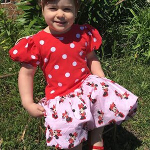 May include: A young girl wearing a red and white polka dot dress with a red bow in her hair. The dress has a pink skirt with a Minnie Mouse print. She is wearing red shoes.