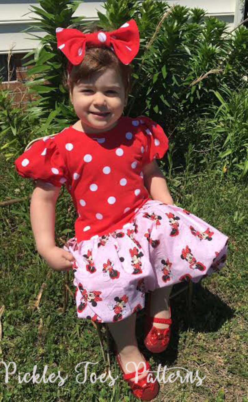 May include: A young girl wearing a red and white polka dot dress with a red bow in her hair. The dress has a pink skirt with a Minnie Mouse print. She is wearing red shoes.