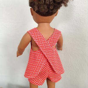 Puede incluir: Un vestido de mu&ntilde;eca rojo y blanco con una correa cruzada en la espalda. El vestido tiene un patr&oacute;n de corazones blancos sobre un fondo rojo. El vestido lo lleva una mu&ntilde;eca con el pelo rizado marr&oacute;n.
